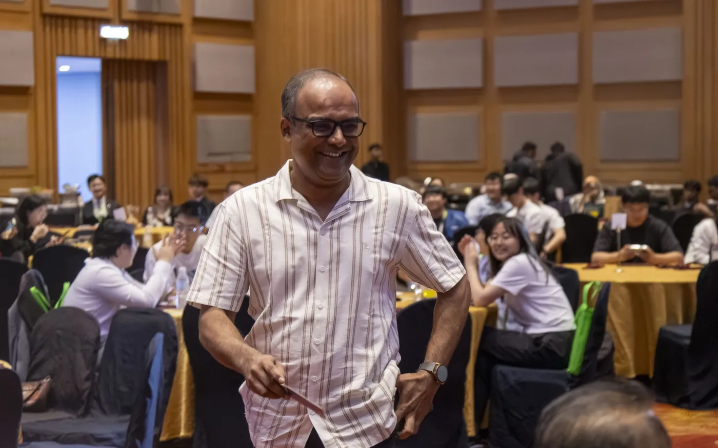 A delegate walking between the tables of a conference hall, with delegates seated at work behind him.