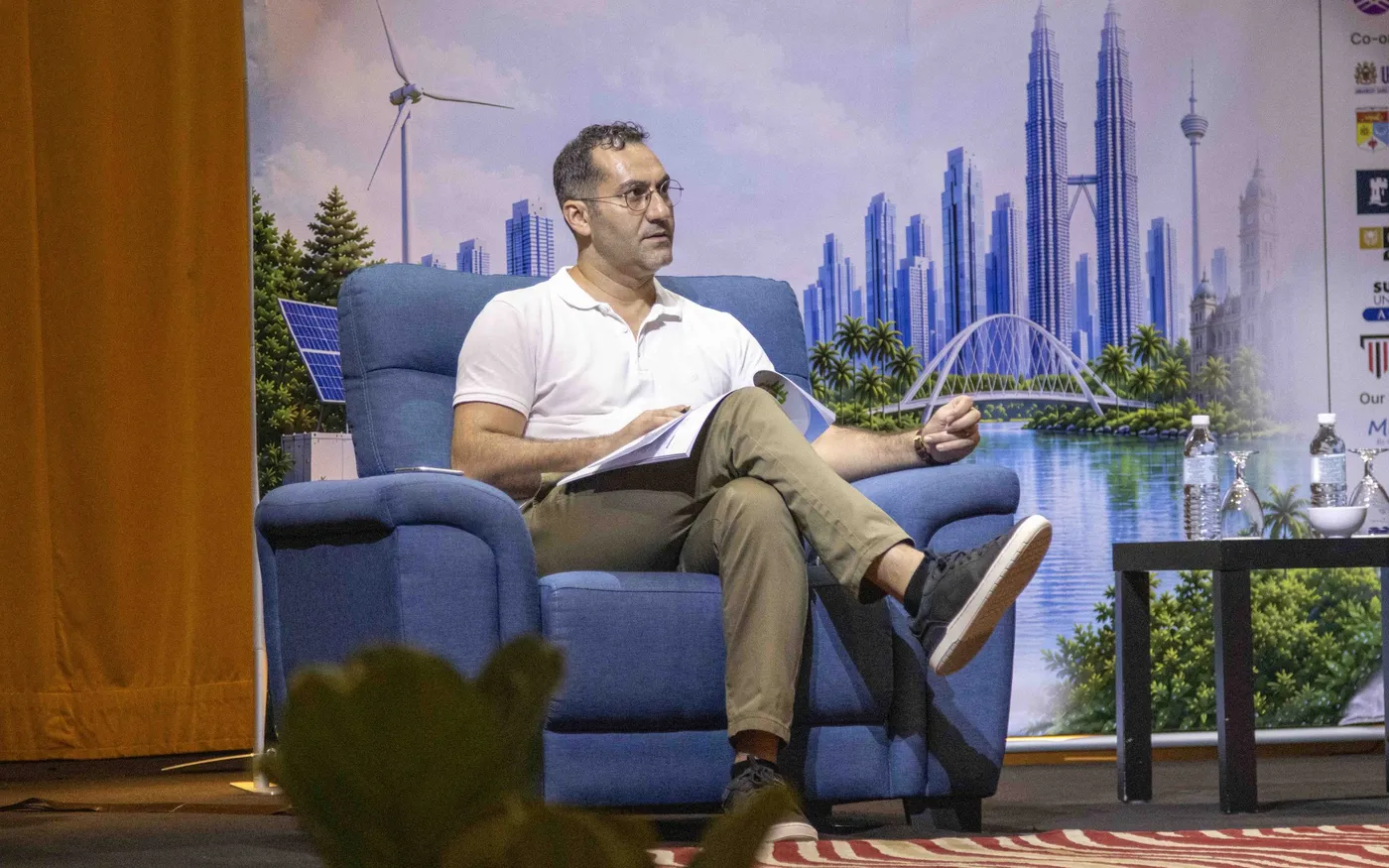 A speaker seated in an armchair on a conference stage, papers in hand, in front of a painted backdrop of a city skyline.