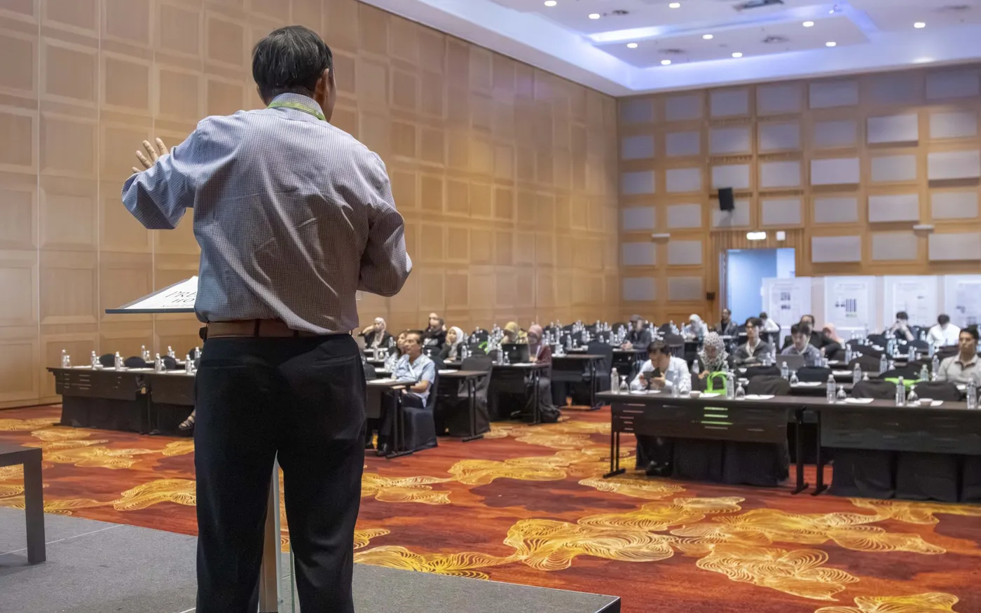 A speaker standing at the front of a conference room, seen from behind, addressing rows of seated delegates.